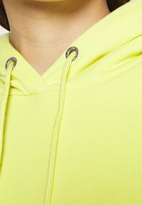 Zbliżenie na osobę w jasnopomarańczowej bluzie z kapturem, z sznurkiem przewleczonym przez srebrne oczka przy dekolcie.