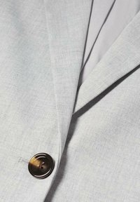 Světle šedý blazer s jedním velkým tmavě hnědým knoflíkem a viditelnými klopami, které ukazují texturu látky a detaily šití.