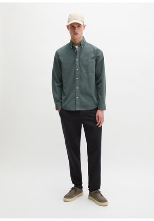 Junger Mann, der ein grünes Hemd mit Knöpfen, schwarze Hose, beige Turnschuhe und eine beige Baseballkappe trägt, steht vor einem weißen Hintergrund.