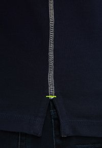 Dark navy t-shirt with a contrasting white stitched seam down the center and a small neon green accent. Features a split hem design.