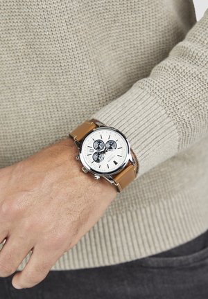 Pols van een man met een beige gebreide trui, een zilveren horloge met een witte wijzerplaat, drie zwarte subdials en een lichtbruine leren band.