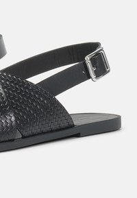 Μαύρη δερμάτινη σάνταλ με πλεγμένη υφή, ρυθμιζόμενο λουράκι στον αστράγαλο με μεταλλική πόρπης και επίπεδη σόλα.