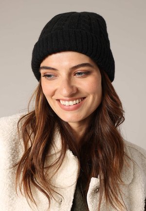 Bonnet en tricot noir avec une texture côtelée, doté un revers replié. Porté avec un manteau texturé de couleur crème et un haut olive en dessous.