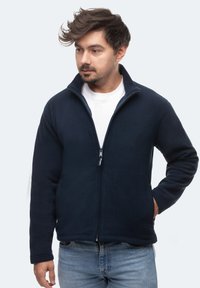 Giacca in pile blu navy con colletto alto e zip a tutta lunghezza. Caratterizzata da maniche lunghe, tasche laterali e una texture morbida.
