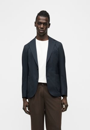 Άνδρας που φοράει σκούρο μπλε σακάκι, λευκό μπλουζάκι με στρογγυλή λαιμόκοψη και καφέ παντελόνι, στέκεται μπροστά σε απλό φωτεινό φόντο.