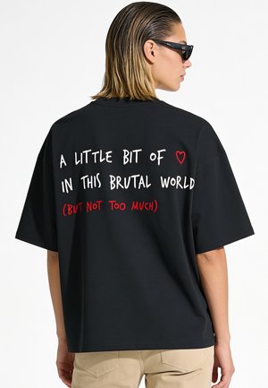 Persona che indossa una maglietta nera oversize con il testo "A little bit of heart in this brutal world (but not too much)" in lettere bianche e rosse.