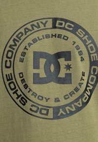 Olivgrünes Baumwoll-T-Shirt mit einem großen schwarzen Logoschema und kreisförmigem Text, der "DC Shoe Company, Established 1994" enthält.