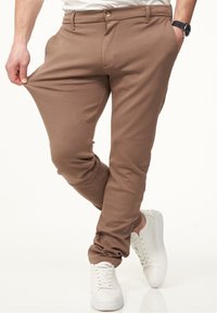 Brown stretch pants with a fitted design, featuring front pockets and a button closure, paired with white sneakers. Smooth texture.