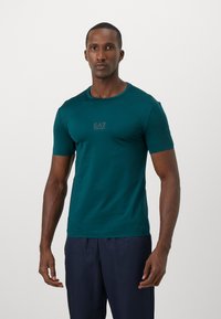 Men's short-sleeve t-shirt in dark teal fabric, featuring a crew neck and a subtle black EA7 logo on the chest. Plain design, close-fitting.