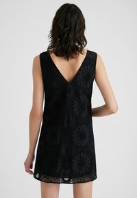 Musta hihaton mekko, jossa on V-pääntie, kukkakuvioinen pitsikuvio ja helma, jossa on hienostunut pitsiyksitys. Rakenteellinen kangas ja tiivistetty muotoilu.