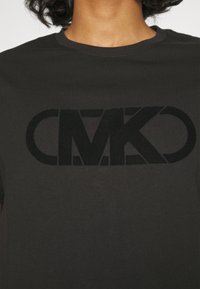 Černé bavlněné tričko s velkým vyraženým logem "CMK" na přední straně. Materiál má hladkou texturu a klasický kulatý výstřih.