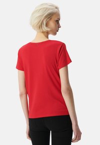 Camiseta roja de manga corta con cuello redondo y una sutil etiqueta de logo en la parte posterior, hecha de una tela suave y ligera. Combinada con pantalones negros.