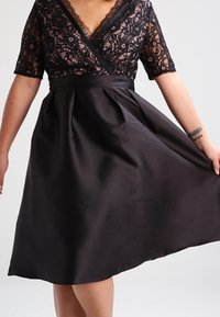 Svart klänning med en spetslivstycke, korta ärmar, V-ringning och en plisserad satängkjol som skapar en figurnära midja och en flödande silhuett.