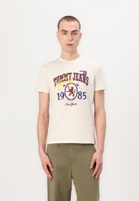 Bežs kokvilnas T-krekls ar grafisku apdari sarkanā, zilā un dzeltenā krāsā, uz kura ir uzraksts "TOMMY JEANS 1985 Ņujorka". Īsās piedurknes un apaļa kakla dizains.