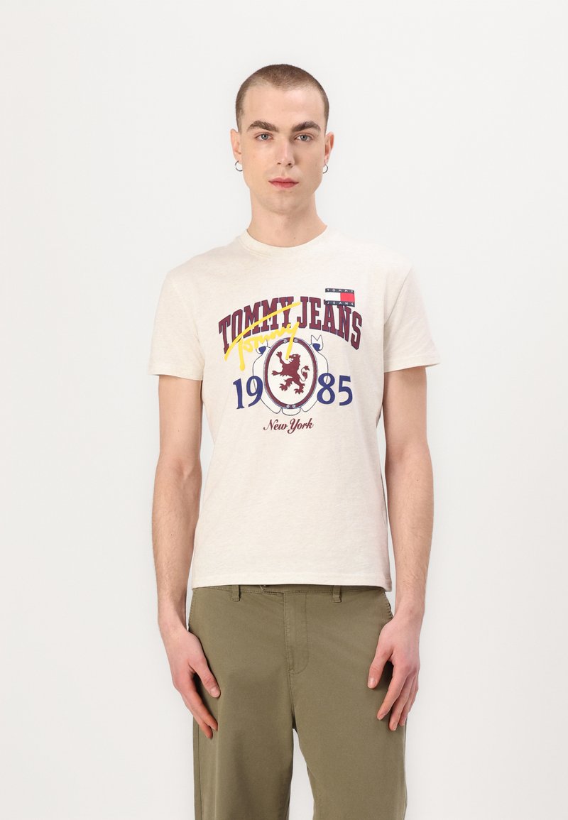 Bežs kokvilnas T-krekls ar grafisku apdari sarkanā, zilā un dzeltenā krāsā, uz kura ir uzraksts "TOMMY JEANS 1985 Ņujorka". Īsās piedurknes un apaļa kakla dizains.