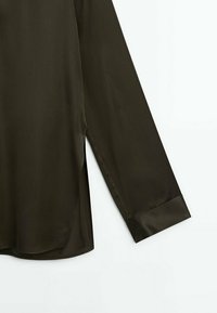Manche de chemise olive foncé à manches longues avec poignet boutonné et fente de couture latérale sur fond blanc.