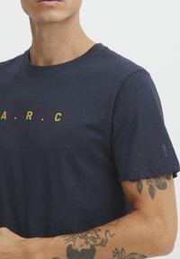 Tamsiai mėlynos spalvos medvilnės marškinėliai su geltonu „A.R.C.“ tekstu, trumpomis rankovėmis ir subtiliu akcentu ant rankogalio. Lygus paviršius.