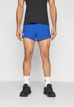 Homme debout portant un short de sport bleu, des chaussettes blanches Adidas, des chaussures de course noires et un t-shirt noir sur un fond uni.
