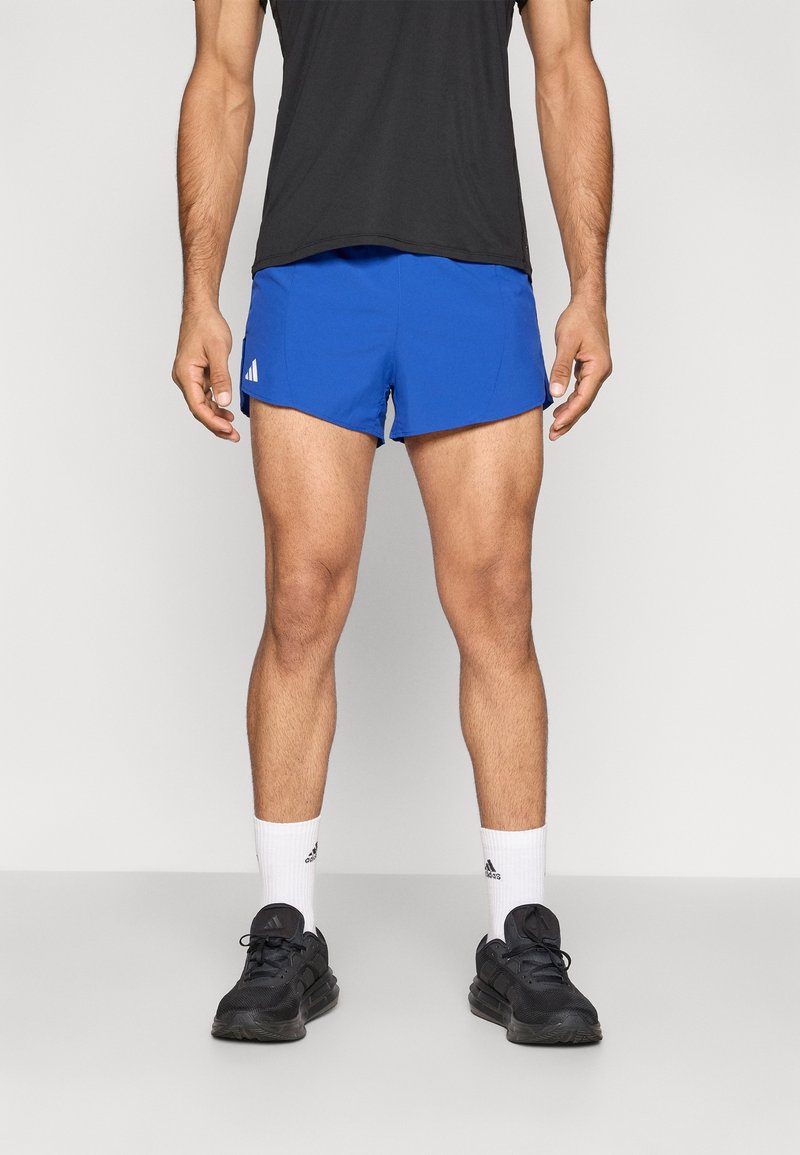 Homme debout portant un short de sport bleu, des chaussettes blanches Adidas, des chaussures de course noires et un t-shirt noir sur un fond uni.