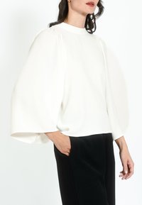 Blusa bianca a maniche lunghe con spalle a palloncino e collo alto, abbinata a pantaloni neri su misura. Tessuto liscio, design minimalista.