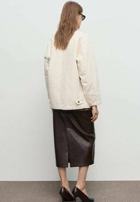 Lichtbeige oversized jas met lange mouwen, gecombineerd met een donkerbruine leren rok met een split aan de achterkant. Zwarte hakken maken de look compleet.