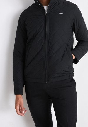 Sort quilted jakke med høj krave, lynlås lukning og sidelommer, prydet med et logo på venstre bryst. Glat materiale, letvægtsdesign.