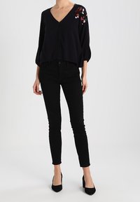 Schwarzes V-Ausschnitt-Top mit gefalteten Ärmeln, das auf einer Schulter mit floraler Stickerei versehen ist. Kombiniert mit schwarzen Skinny-Jeans und schwarzen High Heels.