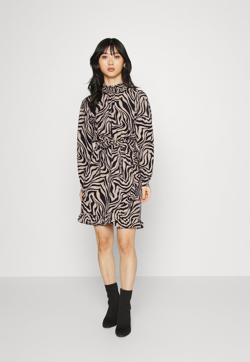 Vestido estampado em bege e padrão de zebra preto, com bainha com babados e cintura amarrada. Estilizado com botas pretas de tornozelo.