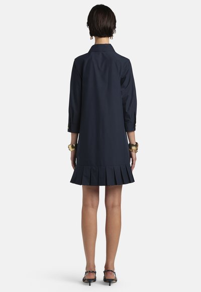 Žena stojaca chrbtom, oblečená v tmavomodrých šatách s dlhým rukávom a riaseným lemom, v čiernych sandáloch na podpätku, doplnená zlatými náramkami.