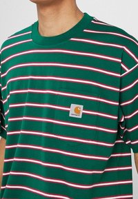 Grønstribet t-shirt med hvide og røde vandrette striber, som har en brystlomme og et firkantet logo-patch. Korte ærmer, afslappet fit.