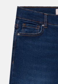 Jeans de ganga azul escuro com costura laranja, apresentando um pequeno bolso frontal e um botão metálico. Textura suave com um design ajustado.