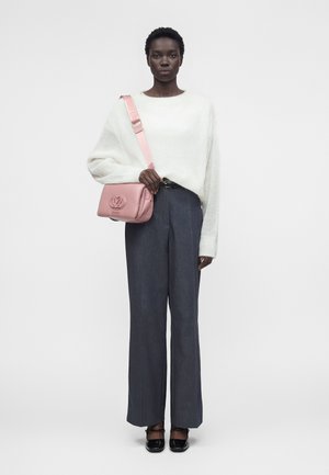 Kvinne iført en hvit, lodden genser, vide mørkegrå bukser, svarte sko og bærer en rosa skulderveske med stor logo.