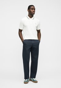 Άνδρας όρθιος με τα χέρια στις τσέπες, φοράει λευκό κοντομάνικο πόλο, ναυτικό μπλε παντελόνι και πράσινα αθλητικά παπούτσια, μπροστά από απλό φόντο.