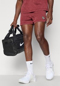 Bolsa deportiva negra con el logo blanco de Nike, pantalones cortos rojos, zapatillas deportivas blancas y calcetines blancos con detalles negros, exhibidos sobre un fondo neutro.