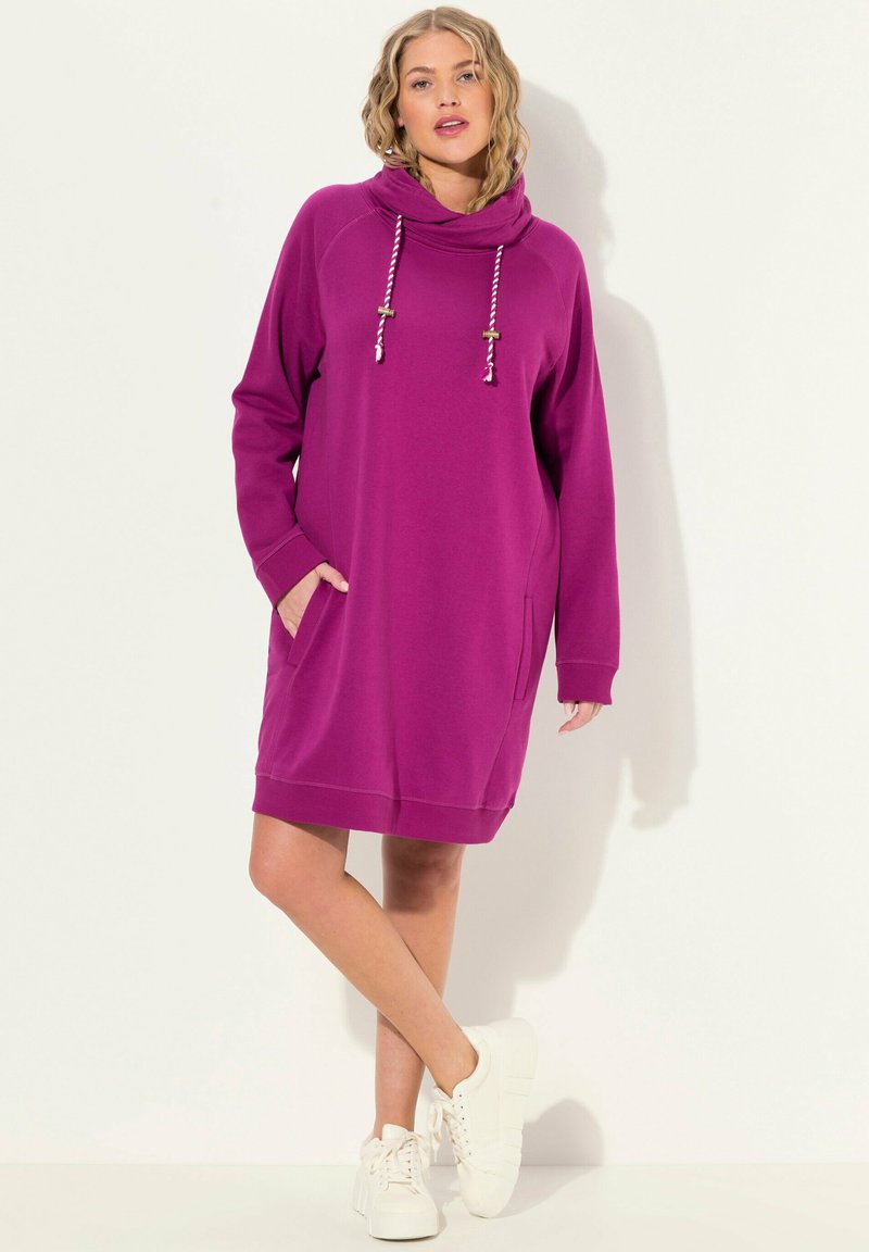 Robe à capuche violette avec une coupe décontractée, dotée de poches latérales, d'une capuche avec cordon de serrage et d'un ourlet côtelé. Associée à des baskets blanches.