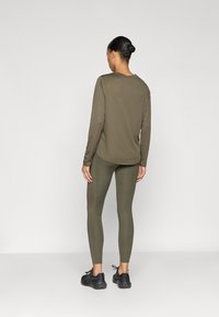 Λαδί μακρυμάνικο πουκάμισο και κολάν από απαλό ύφασμα, σε συνδυασμό με μαύρα αθλητικά παπούτσια, απεικονιζόμενα από πίσω.