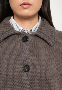 Cappotto lavorato a maglia marrone con un motivo testurizzato, dotato di due bottoni neri e cuciture blu lungo il bordo del colletto. Una camicia a righe è visibile sotto.