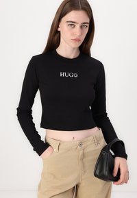 Top cropped nero a maniche lunghe con scollatura rotonda e testo "HUGO" bianco con grafica di smiley. Abbinato a pantaloni beige e una piccola borsa nera.