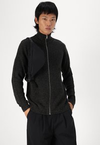 Schwarzer Strickzipper mit vertikaler Rippung, hohem Kragen und langen Ärmeln, kombiniert mit einer schwarzen Umhängetasche.