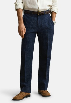 Homme portant un pantalon bleu marine texturé, une ceinture tressée marron, une chemise blanche aux manches retroussées, des mocassins marron et une montre-bracelet argentée, debout les mains dans les poches.