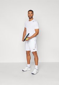 Maglietta atletica bianca a maniche corte e pantaloni shorts, con righe laterali e logo. Il modello tiene una racchetta da tennis e una palla, indossando scarpe da ginnastica bianche.