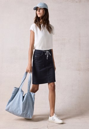 Jeanskjol med fickor, mörkblå färg, kombinerad med en vit t-shirt, ljusblå keps och en stor ljusblå tygväska. Vita sneakers.