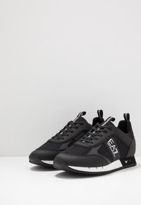 Sneaker sportive nere con pannelli in rete, lacci piatti e punta rinforzata. Presentano un logo bianco e una suola a contrasto per stabilità.
