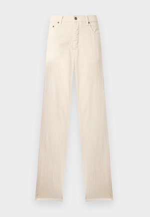 Jeune homme debout portant un polo noir à manches longues, un pantalon beige à jambes larges et des baskets blanches sur un fond blanc uni.