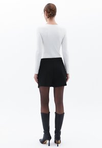 Witte shirt met lange mouwen, zwarte op maat gemaakte shorts, doorzichtige zwarte panty’s en kniehoge zwarte stilettoboots; achteraanzicht, eenvoudig ontwerp.
