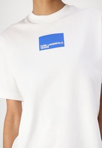 Tricou alb din bumbac, cu un decolteu rotund și mâneci scurte. Prezintă un logo rectangular albastru cu textul "KARL LAGERFELD JEANS" în alb.