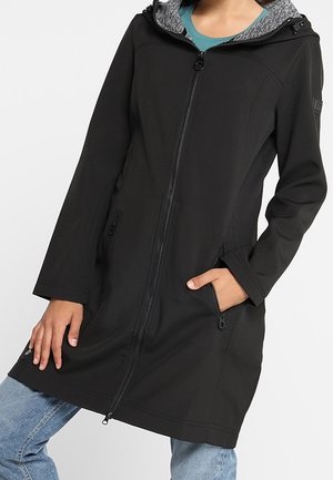 Vrouw draagt een zwarte lange jas met ritssluiting en een grijze voering in de capuchon, handen in de zakken, gecombineerd met lichtblauwe jeans.