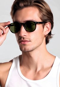 Occhiali da sole in plastica nera con lenti verdi rotonde, indossati da un uomo dalla carnagione chiara. La montatura presenta un motivo tartarugato e braccioli sottili.