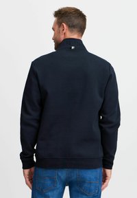 Sudadera azul marino con cuello alto, con un logotipo en relieve en la parte posterior. Tejido suave y texturizado con un corte relajado.