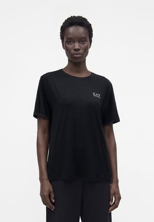 T-shirt noir à manches courtes avec petit logo blanc EA7 Emporio Armani porté par une personne debout sur un fond blanc uni.